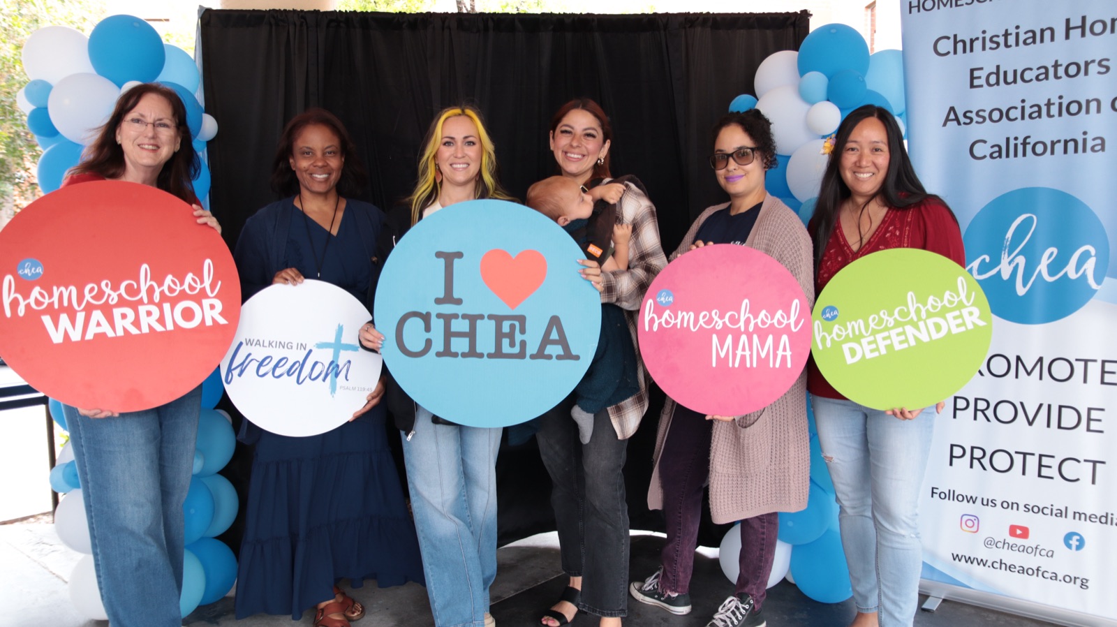 Homeschool moms at a CHEA event holding &lsquo;I love CHEA&rsquo; and &lsquo;Homeschool Mama&rsquo; signs